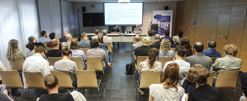 Eine Gruppe von Menschen sitzt in einem modernen Konferenzraum und blickt nach vorne, wo eine Präsentation stattfindet. Zwei Redner sind am Tisch zu sehen, während die Teilnehmer aufmerksam zuhören. Im Hintergrund eine Leinwand mit projiziertem Text. Der Raum hat helle Wände und eine moderne Einrichtung.