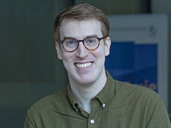 Person indoors in an olive green button-up shirt, with dark blonde hair, standing in front of a blurred background.