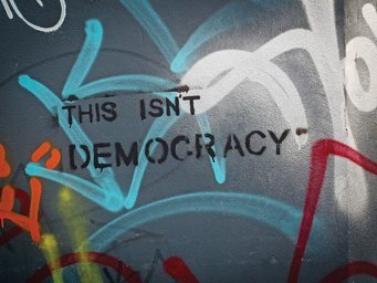 A wall displays graffiti text reading "This isn't democracy" in black. The background is covered with various colorful spray paint tags, including red, blue, and yellow accents.