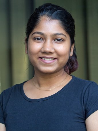 An individual with dark hair, dressed in a navy shirt and a delicate gold necklace, standing in front of a blurred backdrop.