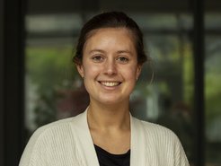 Das Foto zeigt die junge Wissenschaftlerin Pauline Kohlhase, die zur Doktorandensprecherin gewählt wurde. Ihr dunkelblondes Haar hat sie vollständig zum Rücken gekämmt und gebunden, sie strahlt ein ansteckendes Lächeln aus, das Freundlichkeit und Wärme vermittelt. Unter der weißen Jacke schimmert ein schwarzes Hemd unterhalb des frei gelegten Halses heraus.
