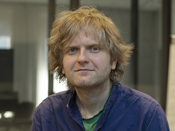 A person with unkempt blonde hair wearing a blue shirt sits in an office setting with glass walls and a flipchart in the background.
