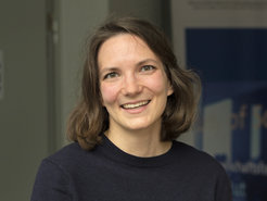 A person with shoulder-length brown hair, wearing a navy sweater, is positioned indoors. In the background, a partially visible poster or sign with blue and white colors is present.