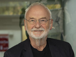 Elderly man with white hair and grey beard, wearing a dark blazer over a black shirt. He is positioned indoors, with blurred background elements including greenery and signage.
