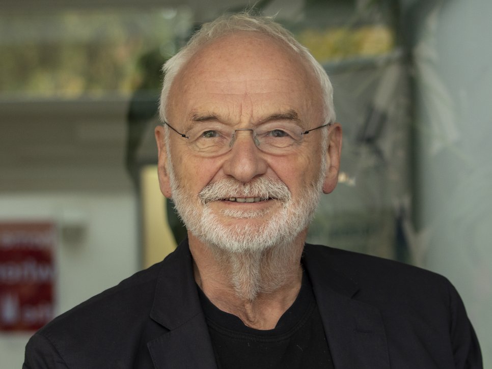 Elderly man with white hair and grey beard, wearing a dark blazer over a black shirt. He is positioned indoors, with blurred background elements including greenery and signage.