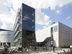 The Copenhagen Business School's modern structure features a glass facade and is surrounded by a large number of parked bicycles, with pedestrians walking nearby on a sunny day.