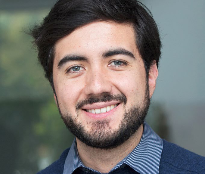 Person in dunkelblauem Hemd und Pullover, Hintergrund zeigt unscharfe graue und grüne Bereiche.