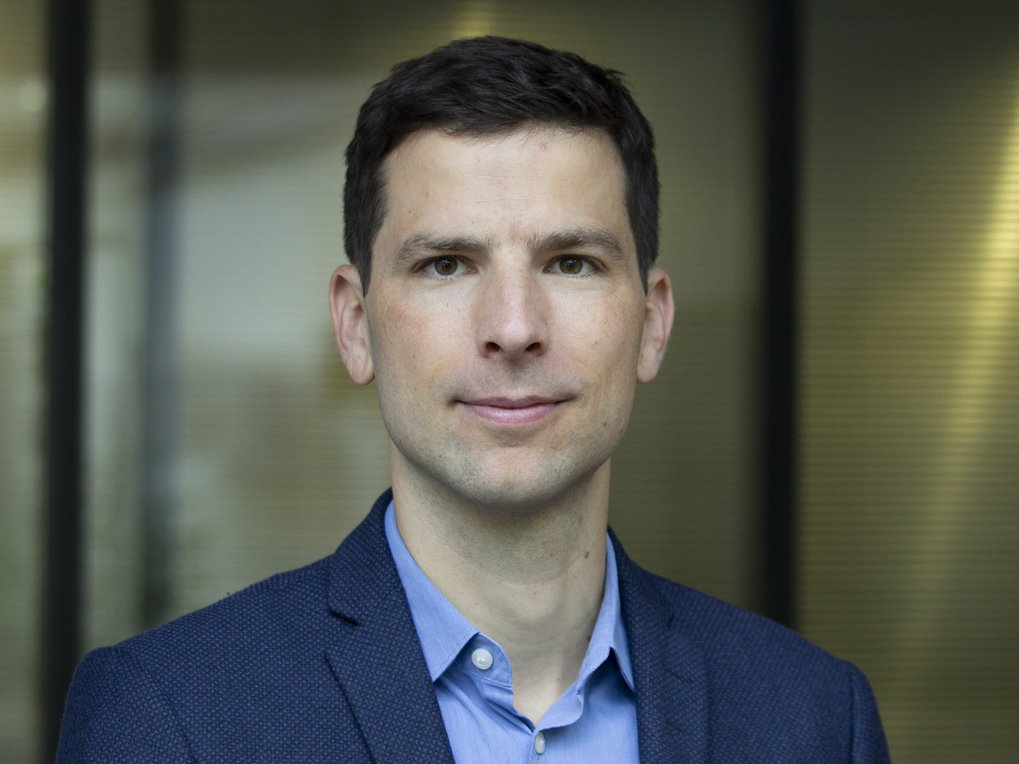 Person in einem strukturierten blauen Sakko über einem hellblauen Hemd. Der Hintergrund ist unscharf, möglicherweise ein modernes Gebäude.