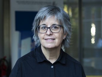 Eine Person mit grauem Haar und Brille trägt ein schwarzes Hemd, steht vor einem Gebäude mit Glasfenstern und einem blauen Plakat im Hintergrund.