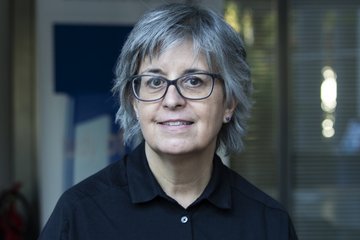 Eine Person mit grauem Haar und Brille trägt ein schwarzes Hemd, steht vor einem Gebäude mit Glasfenstern und einem blauen Plakat im Hintergrund.