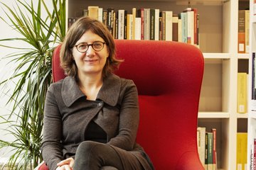 Eine Frau in grauer Kleidung sitzt auf einem roten Sessel. Links im Bild ist eine grüne Zimmerpflanze. Im Hintergrund befindet sich ein Bücherregal mit verschiedenen Büchern.