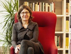 Eine Frau in grauer Kleidung sitzt auf einem roten Sessel. Links im Bild ist eine grüne Zimmerpflanze. Im Hintergrund befindet sich ein Bücherregal mit verschiedenen Büchern.