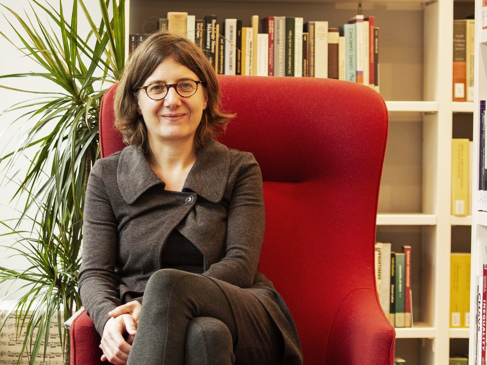 Eine Frau in grauer Kleidung sitzt auf einem roten Sessel. Links im Bild ist eine grüne Zimmerpflanze. Im Hintergrund befindet sich ein Bücherregal mit verschiedenen Büchern.