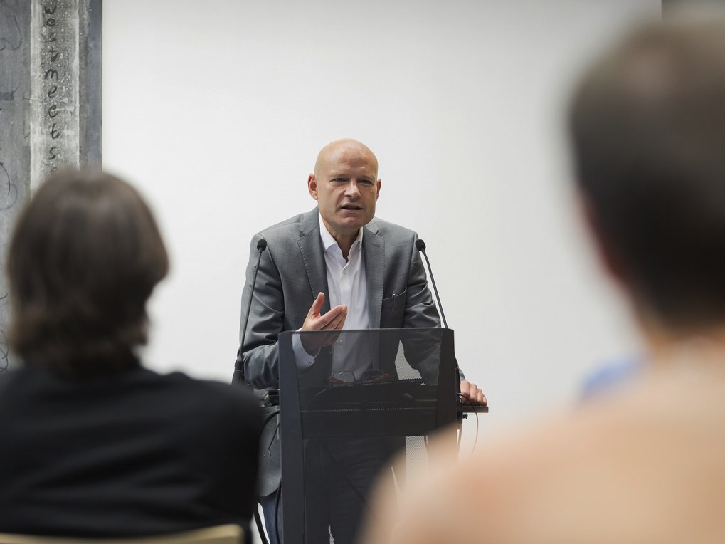 Ein Mann im Anzug spricht während einer Präsentation an einem Podium, im Vordergrund ist das Publikum zu sehen.