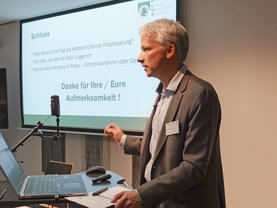 Ein Redner hält vor einem Laptop und einer Leinwand mit deutschem Text eine Präsentation und spricht über Themen im Zusammenhang mit Demokratie und Regierungsführung.