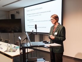 A woman giving a presentation at a conference, standing at a podium with a laptop, while a slide with the title "Today's talk has three parts" is visible behind her.