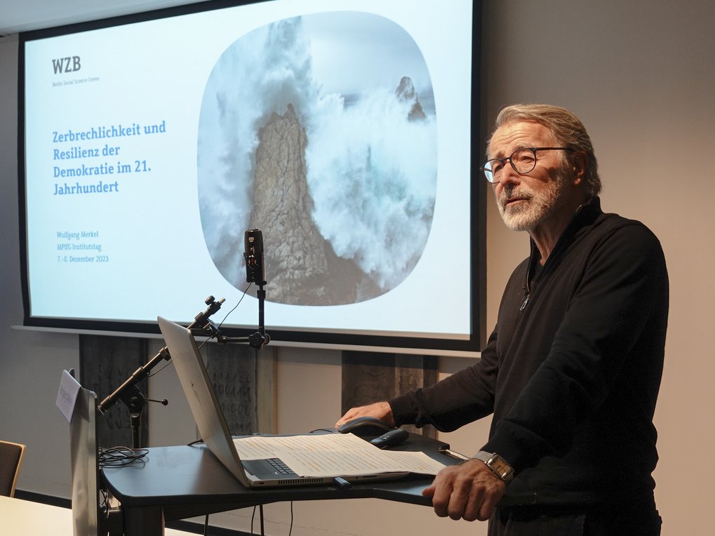 A presenter stands at a podium with a laptop, in front of a slide that discusses the fragility and resilience of democracy in the 21st century.