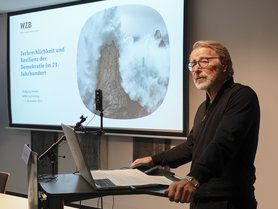 A presenter stands at a podium with a laptop, in front of a slide that discusses the fragility and resilience of democracy in the 21st century.
