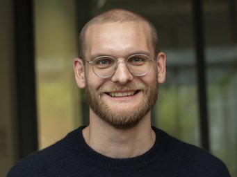 Bald man with a beard wearing a dark sweater, standing in front of a window with green and gray blurred background elements.