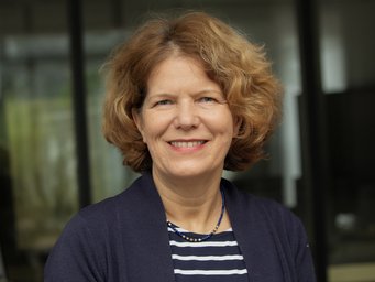 The image features a woman with curly light brown hair, wearing a navy cardigan over a navy and white striped shirt, accessorized with a beaded necklace.