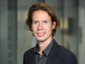 A person with medium-length, wavy hair is standing in front of a blurred background. They are dressed in a dark jacket and grey shirt.