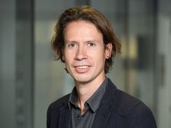 A person with medium-length, wavy hair is standing in front of a blurred background. They are dressed in a dark jacket and grey shirt.