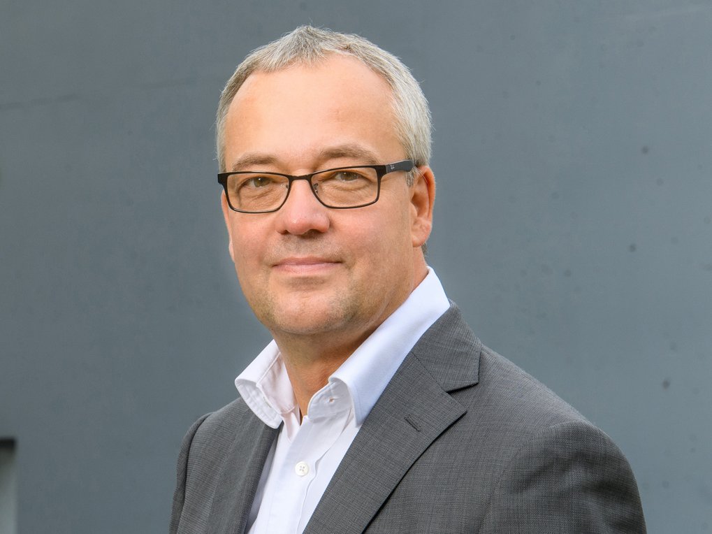 Mittelalterlicher Mann mit grauem Haar, trägt einen dunkelgrauen Anzug und weißes Hemd vor einem schlichten, grauen Hintergrund.