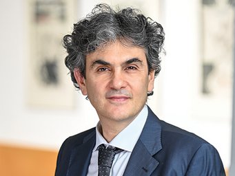 The photo shows Lucio Baccaro, Director at the MPIfG since 2017. In November 2023, he was appointed Honorary Professor at the Institute for Political Science (IFP) and the Institute for Socio-Economic Research (IFSO) at the University of Duisburg-Essen. An individual with curly gray hair is dressed in a dark suit, white shirt, and dotted tie, standing indoors with framed artwork in the background.