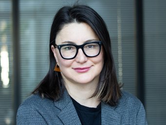 A person with shoulder-length hair is wearing a gray blazer and standing in front of a backdrop of vertical blinds.