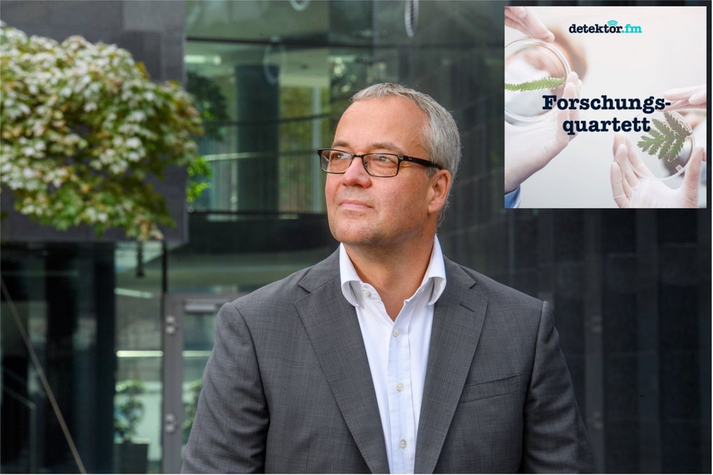 Ein Mann im Anzug steht vor einem modernen Gebäude, mit einem teilweise verdeckten Podcast-Logo, das Hände mit einer Pflanze und den Text „Forschungsquartett” in der Ecke zeigt.