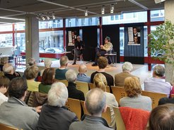 A diverse audience fills a modern venue, listening intently to two men engaged in dialogue on a stage, with promotional posters visible in the background.