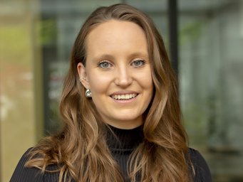 Individual with wavy brown hair, wearing a black turtleneck, positioned in a modern indoor environment with natural light.