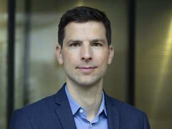 A person is depicted standing in front of a glass wall with a blurred background. The person is wearing a structured blue jacket and light blue shirt.