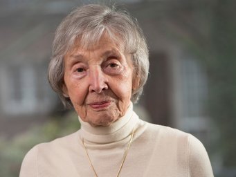 Grey-haired person dressed in a beige turtleneck sweater, adorned with a gold necklace, with a blurred outdoor setting behind.