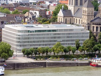 A modern office building is situated by a river, with a row of trees lining the street in front. In the background, a historic church and various residential structures are visible, creating a contrast between contemporary and traditional architecture.