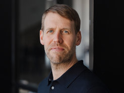 Das Foto zeigt Daniel Mertens, den die Mitgliederversammlung des Vereins der Freunde und Ehemaligen des MPIfG im Dezember 2023 in den Vorstand gewählt hat. Mertens steht schräg zum Ablichter, den Kopf allerdings ihm zugewandt. Kurzes, volles Haar, Ansätze sprießender Barthaare, der Anflug eines Lächelns, das sich noch nicht entschieden hat, das Tageslicht zu sehen - es begegnet dem Betrachter ein ernsthaft gestimmtes Exemplar der Gattung Homo sapiens intellectualis, das wirkt, als hätte man es ungefragt aus der intensiv vertieften Forschungstätigkeit herausgerissen.