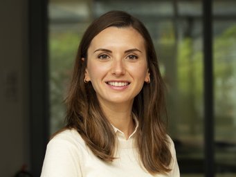 Frau mit schulterlangen braunen Haaren, die ein weißes Oberteil trägt und vor einem großen Glasfenster mit unscharfem Hintergrund steht. Das Foto zeigt Dr. Melike Arslan, die an die Loughborough University wechselt.