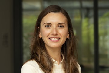 Frau mit schulterlangen braunen Haaren, die ein weißes Oberteil trägt und vor einem großen Glasfenster mit unscharfem Hintergrund steht. Das Foto zeigt Dr. Melike Arslan, die an die Loughborough University wechselt.