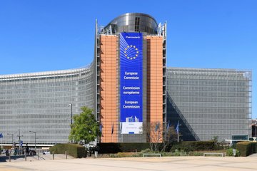 Der Hauptsitz der Europäischen Kommission in Brüssel, an dem ein großes Banner mit dem EU-Emblem und dem Namen der Kommission in Englisch, Französisch und Deutsch angebracht ist. Das Gebäude hat eine moderne Architektur mit Glas- und Stahlelementen und ist von Grünflächen und einem strahlend blauen Himmel umgeben.