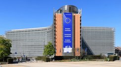 Der Hauptsitz der Europäischen Kommission in Brüssel, an dem ein großes Banner mit dem EU-Emblem und dem Namen der Kommission in Englisch, Französisch und Deutsch angebracht ist. Das Gebäude hat eine moderne Architektur mit Glas- und Stahlelementen und ist von Grünflächen und einem strahlend blauen Himmel umgeben.