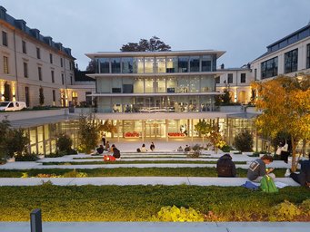 A modern, multi-story building with large glass windows is surrounded by landscaped steps where people are seated, enhanced by warm evening lights.