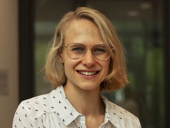 Shoulder-length blonde hair, wearing a white shirt adorned with black crescent shapes, standing indoors against a blurred background featuring green and gray hues.