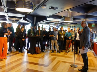 A speaker addresses an attentive audience in a well-lit conference space, accented by geometric ceiling fixtures and parquet flooring. Participants hold drinks and appear engaged.