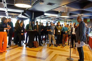 A speaker addresses an attentive audience in a well-lit conference space, accented by geometric ceiling fixtures and parquet flooring. Participants hold drinks and appear engaged.