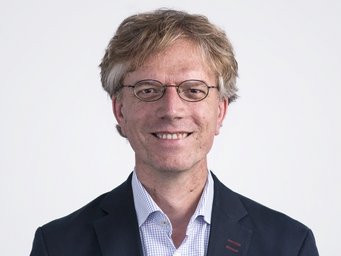 Person dressed in a formal dark blazer with subtle red stitching and a light checkered shirt, having light brown hair.