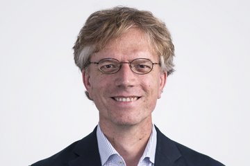 Person dressed in a formal dark blazer with subtle red stitching and a light checkered shirt, having light brown hair.