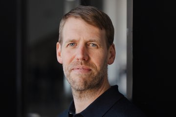 A man with short brown hair and a well-groomed beard is wearing a dark shirt. He is standing against a wall in an indoor area.