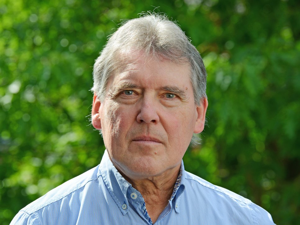 Ein älterer Herr mit grauen Haaren trägt ein blau gestreiftes Hemd. Der Hintergrund besteht aus unscharfem Laubwerk.