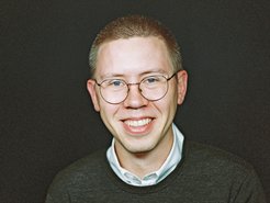 Junges Individuum der Gattung Homo sapiens intellectualis mit braun-grau meliertem Haar, weißem Hemd mit kleinem Kragen, der unter einem dunkelgrauen Pullover hervorsticht. Individuum trägt recht große Brille zur besseren optischen Orientierung in der Umgebugn, es lächelt, ja lacht beinahe, wirkt friedlich-freundlich. Der Hintergrund dagegen ist einfarbig schwarz gehalten und steht dem individuellen Eindruck diametral entgegen.