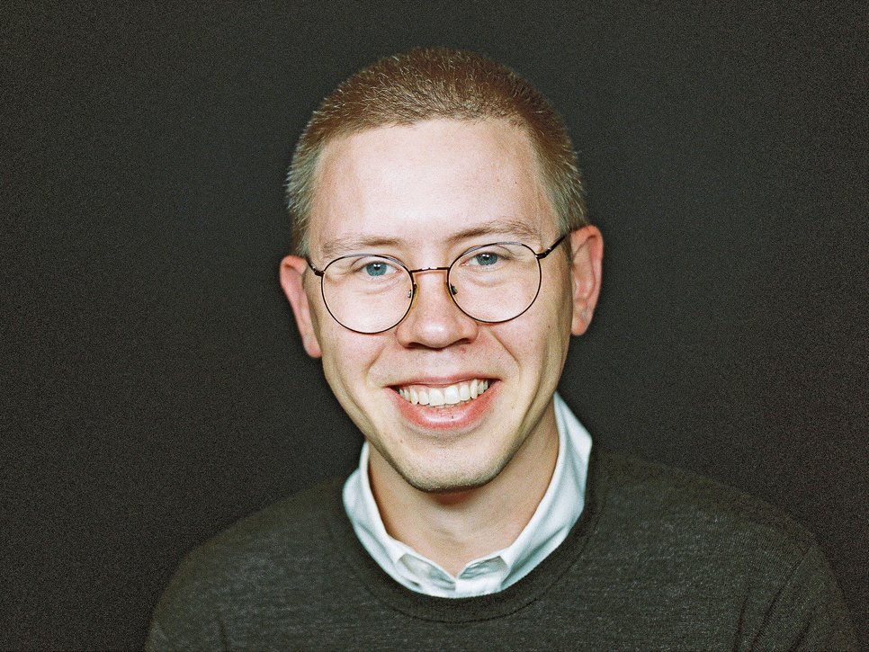 Junges Individuum der Gattung Homo sapiens intellectualis mit braun-grau meliertem Haar, weißem Hemd mit kleinem Kragen, der unter einem dunkelgrauen Pullover hervorsticht. Individuum trägt recht große Brille zur besseren optischen Orientierung in der Umgebugn, es lächelt, ja lacht beinahe, wirkt friedlich-freundlich. Der Hintergrund dagegen ist einfarbig schwarz gehalten und steht dem individuellen Eindruck diametral entgegen.